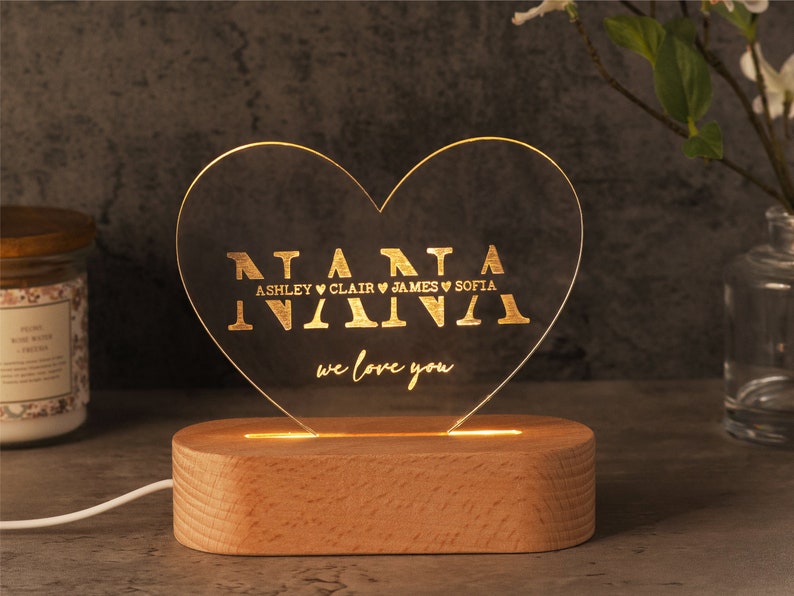 May include: A heart-shaped LED night light with the word "NANA" in gold lettering, along with names and the phrase "we love you." The light sits on a rectangular wooden base. The light is plugged in with a white cord.