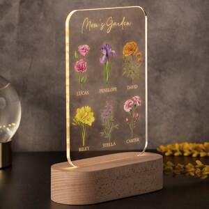 May include: A clear acrylic sign with the words "Mom's Garden" and floral illustrations. The sign features a wooden base with a warm light. The flowers are labeled with names: Lucas, Penelope, David, Riley, Stella, and Carter.