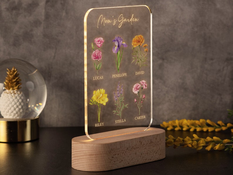 May include: A clear acrylic sign with the words "Mom's Garden" and floral illustrations. The sign features a wooden base with a warm light. The flowers are labeled with names: Lucas, Penelope, David, Riley, Stella, and Carter.
