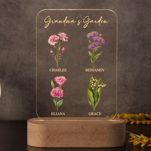 May include: A personalized LED night light with a wooden base. The clear acrylic panel features the words "Grandma's Garden" and illustrations of flowers with names: Charles, Eliana, Benjamin, and Grace. The light is on, illuminating the floral design.