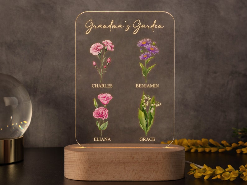 May include: A personalized LED night light with a wooden base. The clear acrylic panel features the words "Grandma's Garden" and illustrations of flowers with names: Charles, Eliana, Benjamin, and Grace. The light is on, illuminating the floral design.