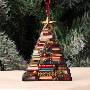 May include: A Christmas tree ornament shaped like a tree made of stacked books. The ornament is decorated with a gold star on top and a red ribbon. The name "HARPER" is printed on the bottom of the ornament.