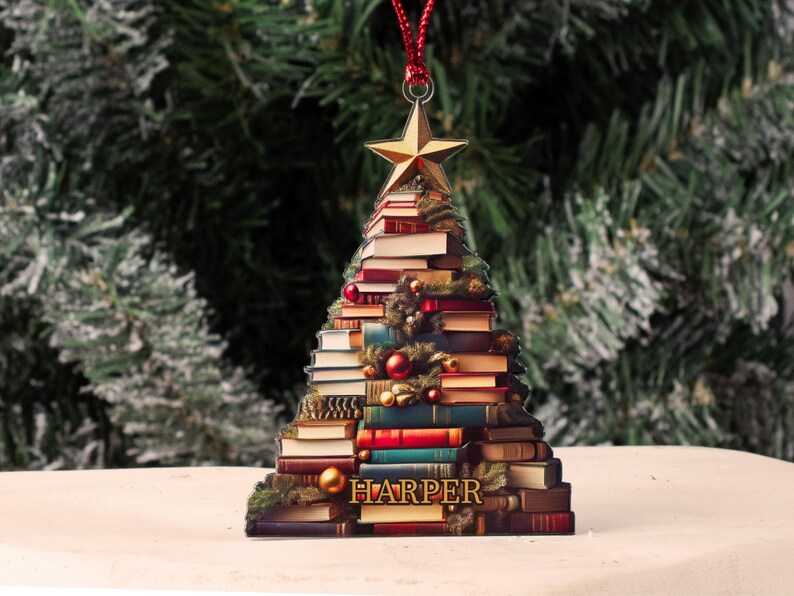 May include: A Christmas tree ornament shaped like a tree made of stacked books. The ornament is decorated with a gold star on top and a red ribbon. The name "HARPER" is printed on the bottom of the ornament.