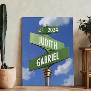 Op de afbeelding: Een groen en wit straatnaambord met de tekst "EST. 2024" op het bovenste bord, "JUDITH" op het middelste bord en "GABRIEL" op het onderste bord. De borden zijn tegen een blauwe lucht met witte wolken.
