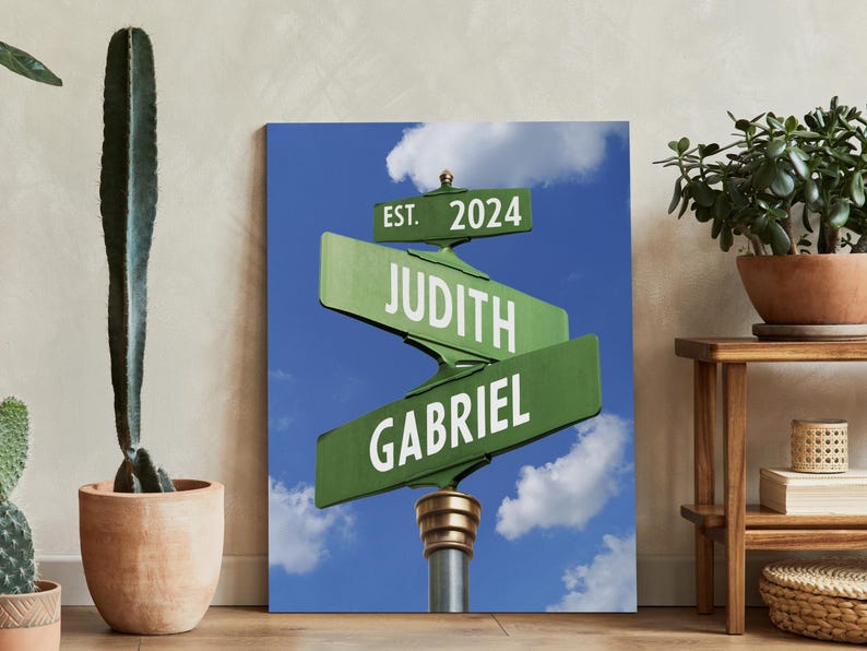 Op de afbeelding: Een groen en wit straatnaambord met de tekst "EST. 2024" op het bovenste bord, "JUDITH" op het middelste bord en "GABRIEL" op het onderste bord. De borden zijn tegen een blauwe lucht met witte wolken.