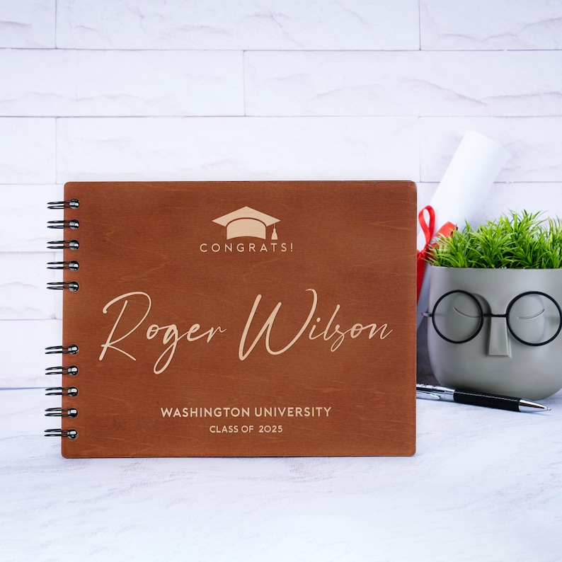 May include: A brown wooden photo album with a gold graduation cap and the text "Congrats!"  The album has a spiral binding and the name "Roger Wilson" is engraved on the front. The text "Washington University Class of 2023" is also engraved on the front.