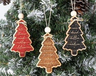 May include: Three wooden Christmas tree ornaments in red, brown, and black. Each ornament features white script names, small dots, and the year 2026 at the base. They are topped with a small wooden bead and a white string for hanging on a tree.