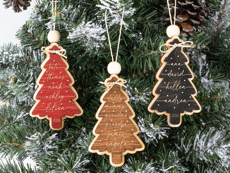 May include: Three wooden Christmas tree ornaments in red, brown, and black. Each ornament features white script names, small dots, and the year 2026 at the base. They are topped with a small wooden bead and a white string for hanging on a tree.