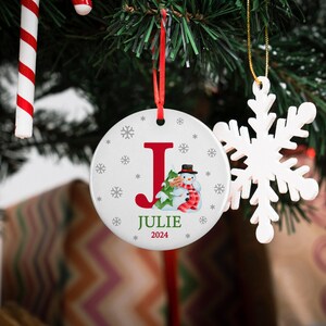 May include: White ceramic Christmas ornament with a red letter J, a snowman holding a small tree, and the name Julie and the year 2024 printed on it.