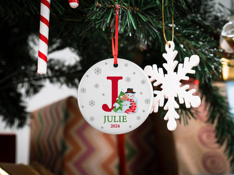 May include: White ceramic Christmas ornament with a red letter J, a snowman holding a small tree, and the name Julie and the year 2024 printed on it.