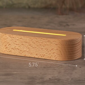 May include: A wooden LED light base, oval-shaped, with a warm yellow glow emanating from a top slot. The base is 5.75 inches long and 1.38 inches high, with a natural wood grain finish. A white cord extends from the base.