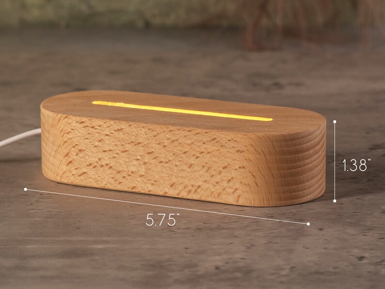 May include: A wooden LED light base, oval-shaped, with a warm yellow glow emanating from a top slot. The base is 5.75 inches long and 1.38 inches high, with a natural wood grain finish. A white cord extends from the base.