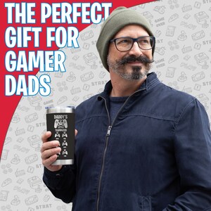 May include: A black stainless steel tumbler with a white design that says "Daddy's Little Teammates" and features a video game controller graphic. The tumbler is being held by a person wearing a blue jacket and a beanie.