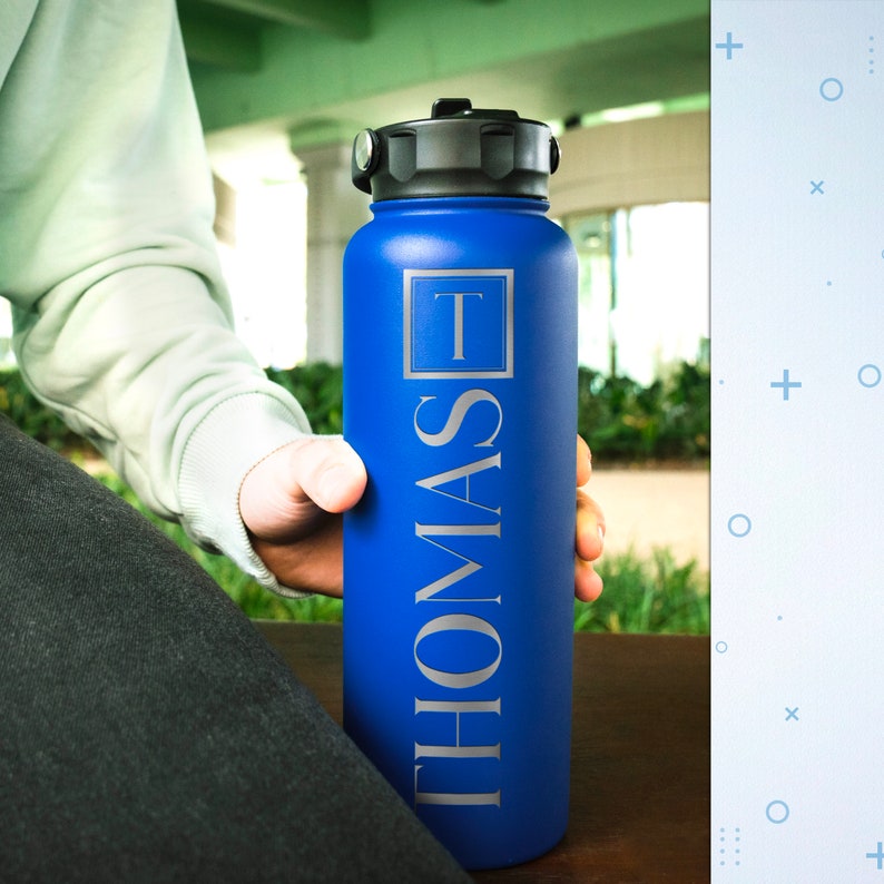 K&ouml;nnte beinhalten: Eine blaue Edelstahl-Wasserflasche mit schwarzem Deckel und einer silbernen Aufschrift "THOMAS" mit einem "T" in einem Quadrat.