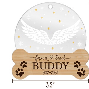 May include: A circular ornament with a bone-shaped wooden base measuring 3.5 inches by 3.5 inches. The top features white angel wings, gold stars, and snowflakes. The base displays the text "forever loved", "BUDDY", and "2012-2023" with paw prints.