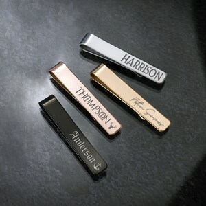 May include: Four different tie clips with personalized engravings. The tie clips are made of silver, gold, and black metal. The engravings include the names "Harrison", "Thompson", "Anderson", and a handwritten signature.