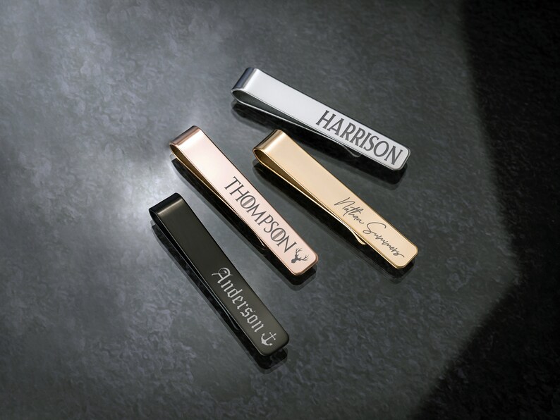 May include: Four different tie clips with personalized engravings. The tie clips are made of silver, gold, and black metal. The engravings include the names "Harrison", "Thompson", "Anderson", and a handwritten signature.