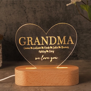 May include: A heart-shaped LED night light with the word "GRANDMA" in gold lettering. Below, names are listed, and the phrase "we love you" is written. The light sits on a wooden base and is illuminated.