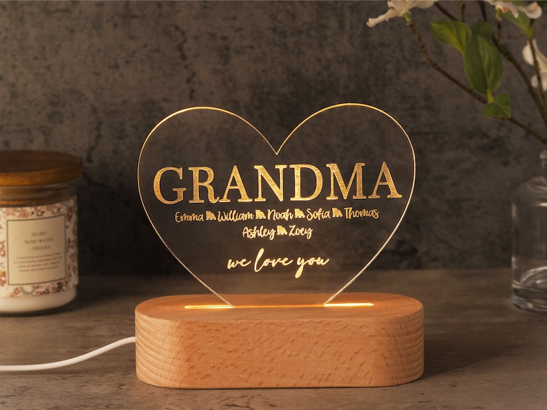 May include: A heart-shaped LED night light with the word "GRANDMA" in gold lettering. Below, names are listed, and the phrase "we love you" is written. The light sits on a wooden base and is illuminated.