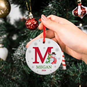 May include: White ceramic Christmas ornament with a red letter M, a snowman, and a green Christmas tree. The ornament also has the name Megan and the year 2024.