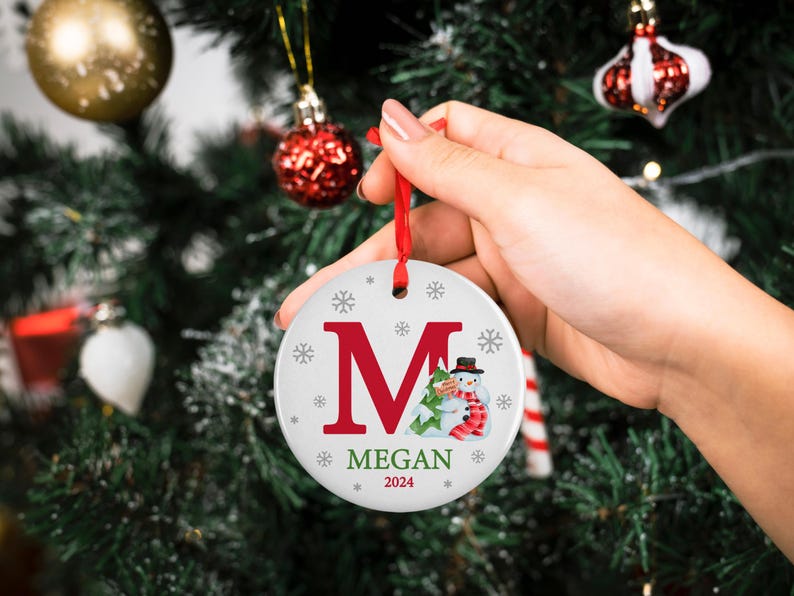 May include: White ceramic Christmas ornament with a red letter M, a snowman, and a green Christmas tree. The ornament also has the name Megan and the year 2024.