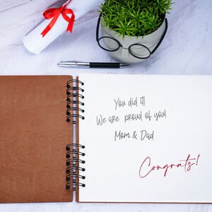 May include: An open brown leather bound journal with a white page showing a handwritten message that says "You did it! We are proud of you! Mom & Dad Congrats!"
