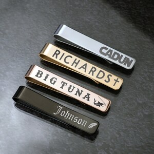 May include: Four different tie clips with engraved names. The tie clips are silver, gold, rose gold, and black.