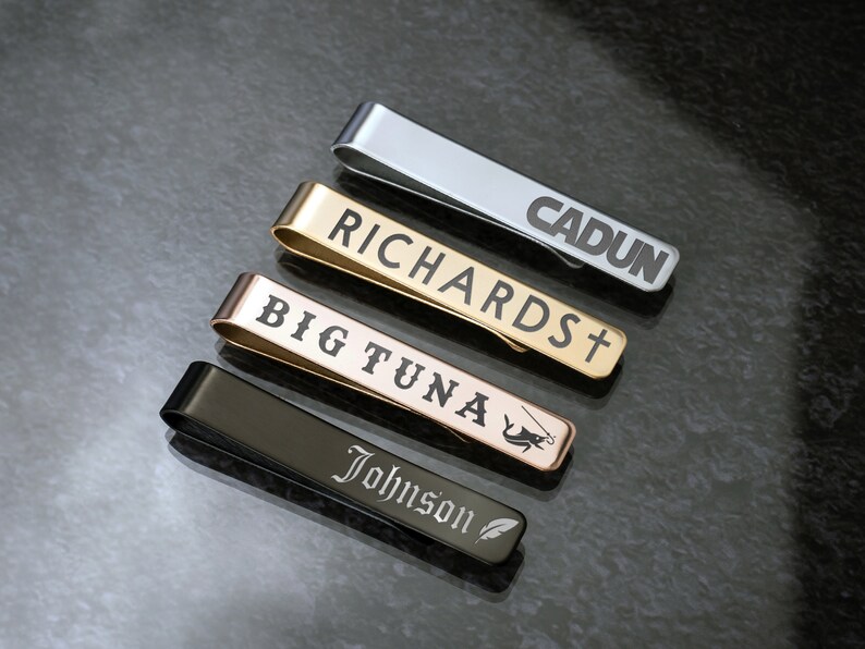 May include: Four different tie clips with engraved names. The tie clips are silver, gold, rose gold, and black.