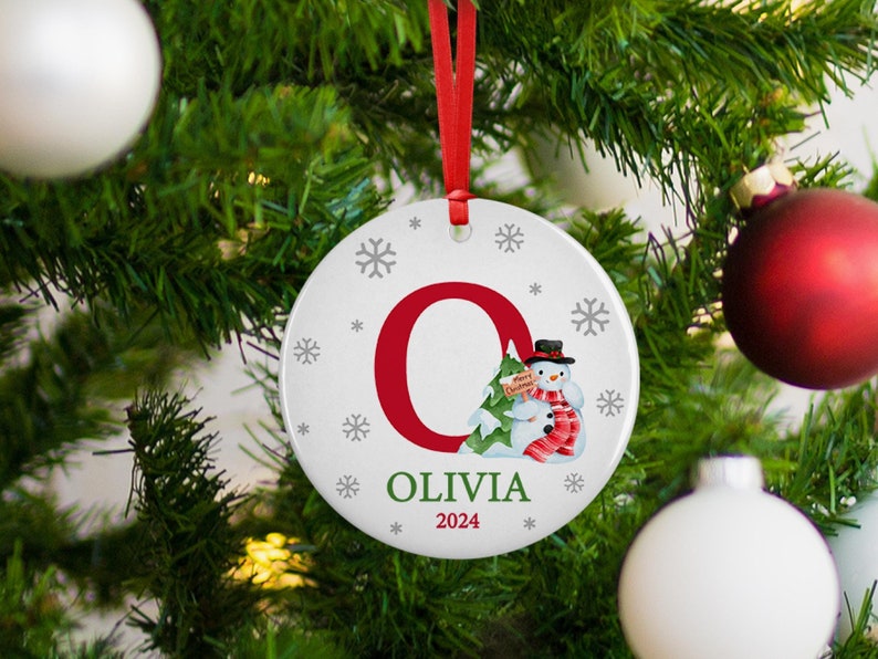May include: White ceramic Christmas ornament with a red letter "O" and a snowman holding a small Christmas tree. The ornament also has the name "Olivia" and the year "2024" printed on it.