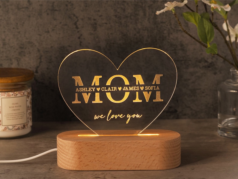 May include: A heart-shaped LED night light with the word "MOM" and the names Ashley, Clair, James, and Sofia. The light is on a wooden base and says "we love you".