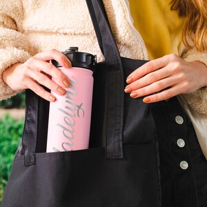 K&ouml;nnte beinhalten: Eine rosa Wasserflasche mit schwarzem Deckel und einer silbernen Aufschrift "Madelyn". Die Flasche befindet sich in einer schwarzen Tragetasche mit einer Jeans-Tasche.