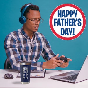 May include: A blue and white stainless steel tumbler with the text "Daddy's Little Teammates" and a graphic of video game controllers. The tumbler is sitting on a desk with a laptop and a video game controller.