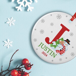 May include: White round Christmas ornament with a red letter J, a snowman holding a sign that says "Merry Christmas", and the name "Justin 2024" in green text. The ornament is decorated with gray snowflakes.