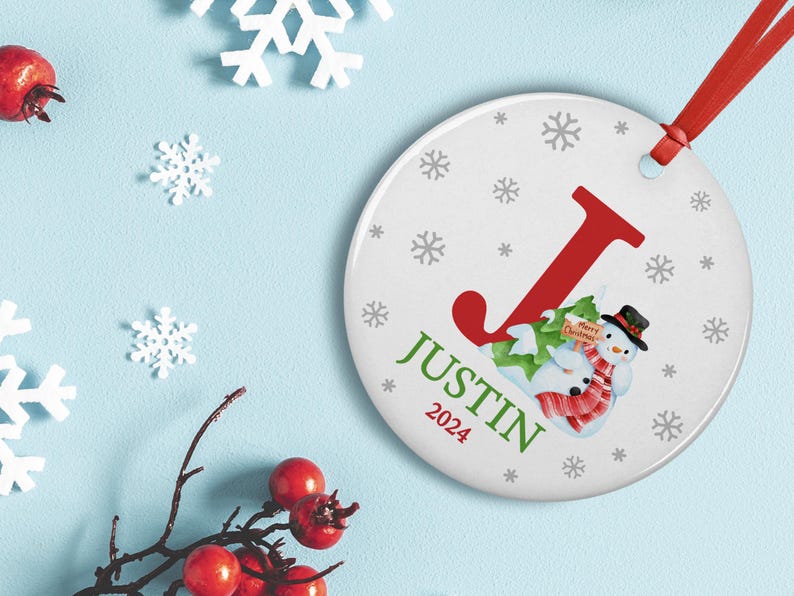 May include: White round Christmas ornament with a red letter J, a snowman holding a sign that says "Merry Christmas", and the name "Justin 2024" in green text. The ornament is decorated with gray snowflakes.