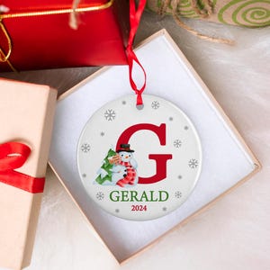 May include: A white round Christmas ornament with a red letter G, a snowman holding a small green tree, and the name "GERALD" with the year "2024" below.