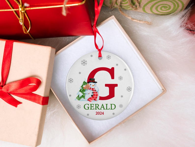May include: A white round Christmas ornament with a red letter G, a snowman holding a small green tree, and the name "GERALD" with the year "2024" below.
