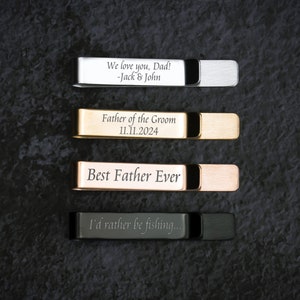 May include: Four engraved metal tie clips in different colors: silver, gold, rose gold, and black. The engravings include "We love you, Dad! -Jack & John", "Father of the Groom 11.11.2024", "Best Father Ever", and "I'd rather be fishing..."