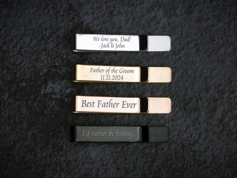 May include: Four engraved metal tie clips in different colors: silver, gold, rose gold, and black. The engravings include "We love you, Dad! -Jack & John", "Father of the Groom 11.11.2024", "Best Father Ever", and "I'd rather be fishing..."