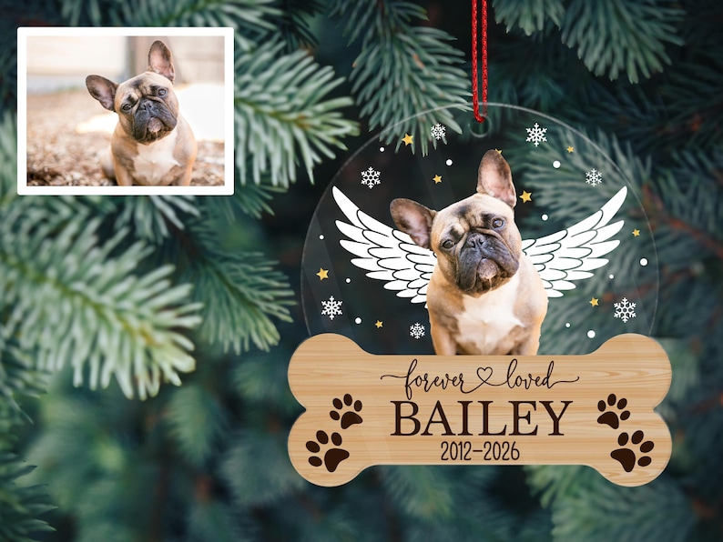 May include: A circular clear acrylic ornament featuring a photo of a fawn French Bulldog with white angel wings, surrounded by small white snowflakes and gold stars. Below, a wooden bone-shaped plaque reads forever loved BAILEY 2012-2026.