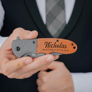 May include: A personalized pocket knife with a wooden handle. The handle is engraved with the name "Nicholas" and the text "Best Man 01.08.2025".