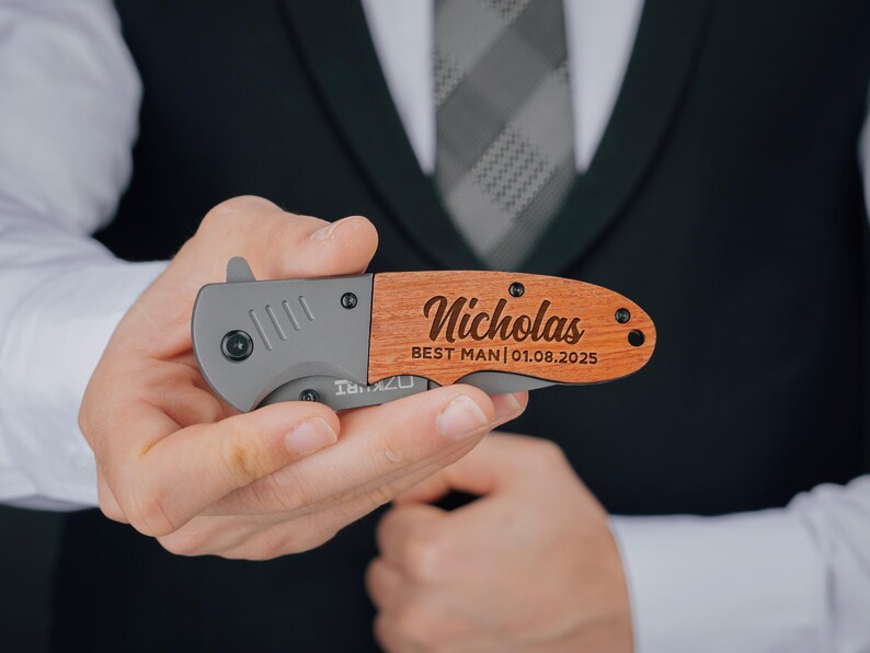 May include: A personalized pocket knife with a wooden handle. The handle is engraved with the name "Nicholas" and the text "Best Man 01.08.2025".