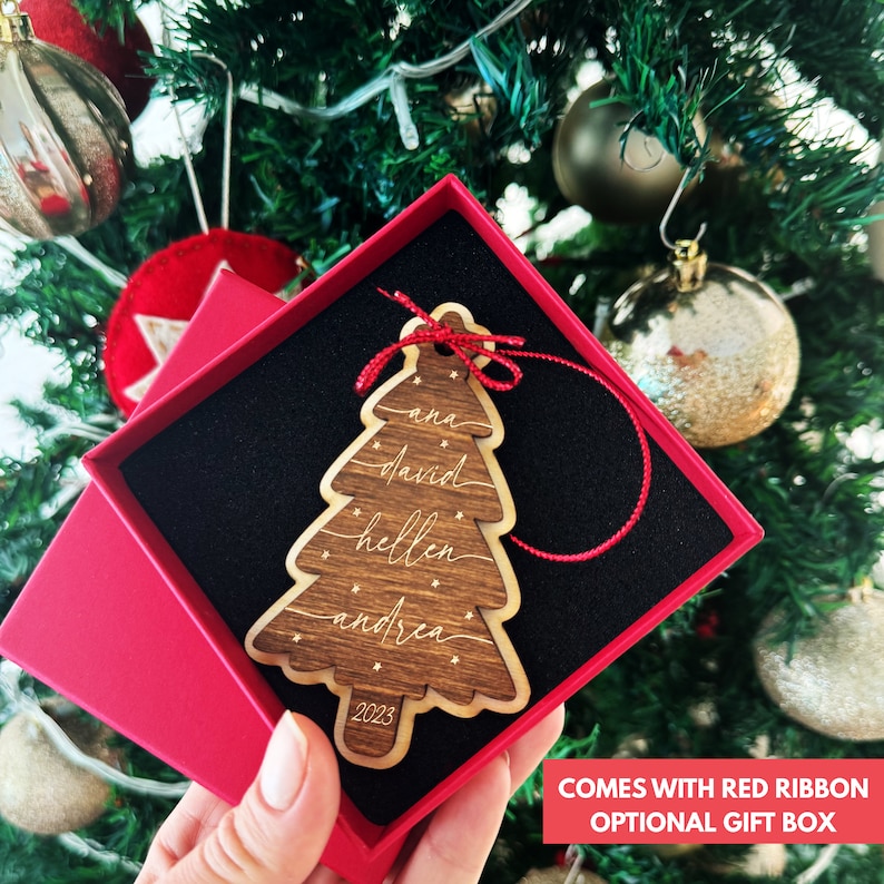 May include: A wooden Christmas tree ornament in a red gift box, featuring the names ana, david, hellen, and andrea engraved in white script, accented with small stars and the year 2023 at the base, finished with a thin red hanging ribbon.