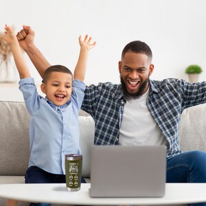 May include: A father and son are cheering while watching a game on a laptop. A green and black stainless steel tumbler with the text "Daddy's Got A Little Terminator" is on the table in front of them.