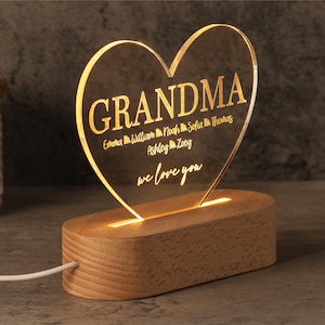 May include: A heart-shaped acrylic LED night light with the word "GRANDMA" and names of loved ones. The light sits on a wooden base and is illuminated with a warm glow. The text "we love you" is also included.