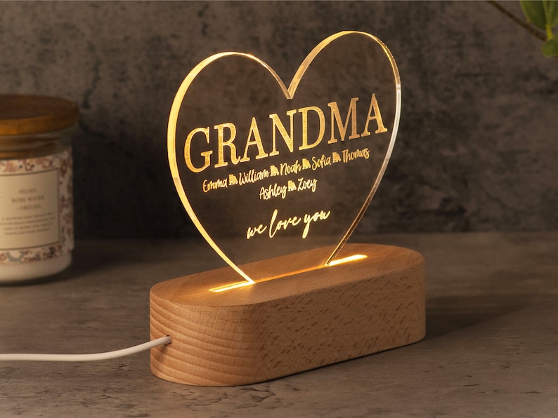 May include: A heart-shaped acrylic LED night light with the word "GRANDMA" and names of loved ones. The light sits on a wooden base and is illuminated with a warm glow. The text "we love you" is also included.
