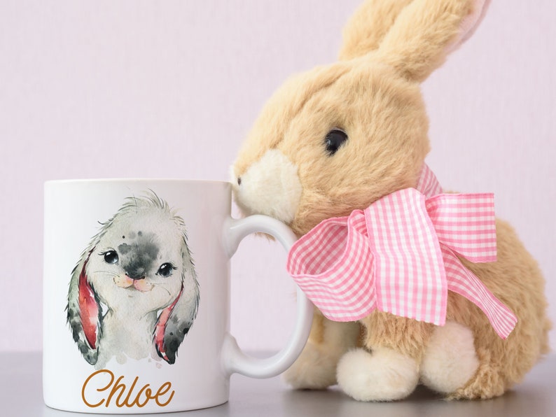 May include: White ceramic mug with a watercolor illustration of a bunny rabbit with the name Chloe printed below the image. A plush brown bunny with a pink gingham bow is leaning against the mug.