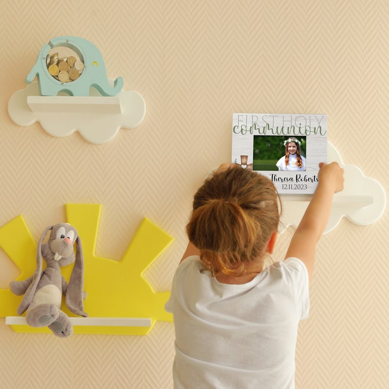 May include: A white cloud-shaped shelf with a picture frame on it. The frame has a white background with the text "FIRST HOLY COMMUNION" in green letters. There is a photo of a girl in the frame. The text "Theresa Roberts 12.11.2023" is below the photo.