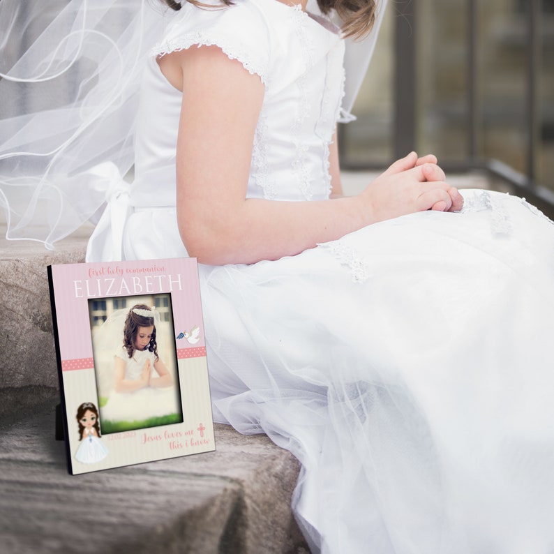 May include: A pink and white picture frame with a photo of a young girl in a white dress. The frame has the text "First Holy Communion ELIZABETH" and "Jesus loves me this I know".