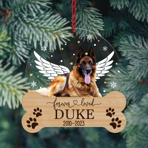 May include: A circular clear acrylic ornament featuring a German Shepherd with white angel wings, surrounded by small stars and snowflakes. Below, a bone-shaped wooden plaque is engraved with the text forever loved DUKE 2010-2023 and paw prints.