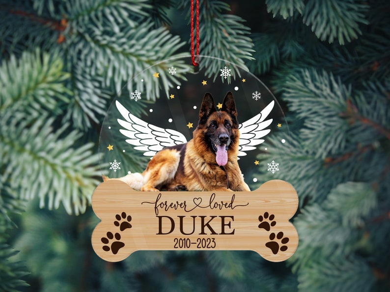 May include: A circular clear acrylic ornament featuring a German Shepherd with white angel wings, surrounded by small stars and snowflakes. Below, a bone-shaped wooden plaque is engraved with the text forever loved DUKE 2010-2023 and paw prints.